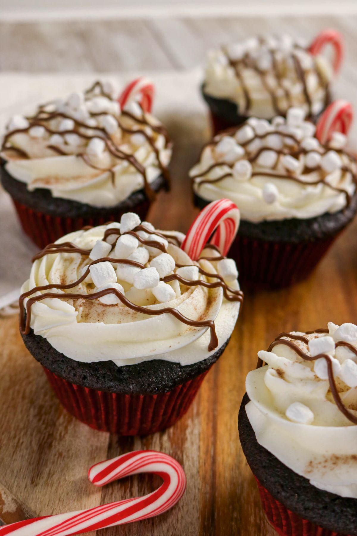Chocolate cupcakes with white frosting, mini marshmallows, chocolate drizzle, and small candy canes, arranged on a wooden surface.