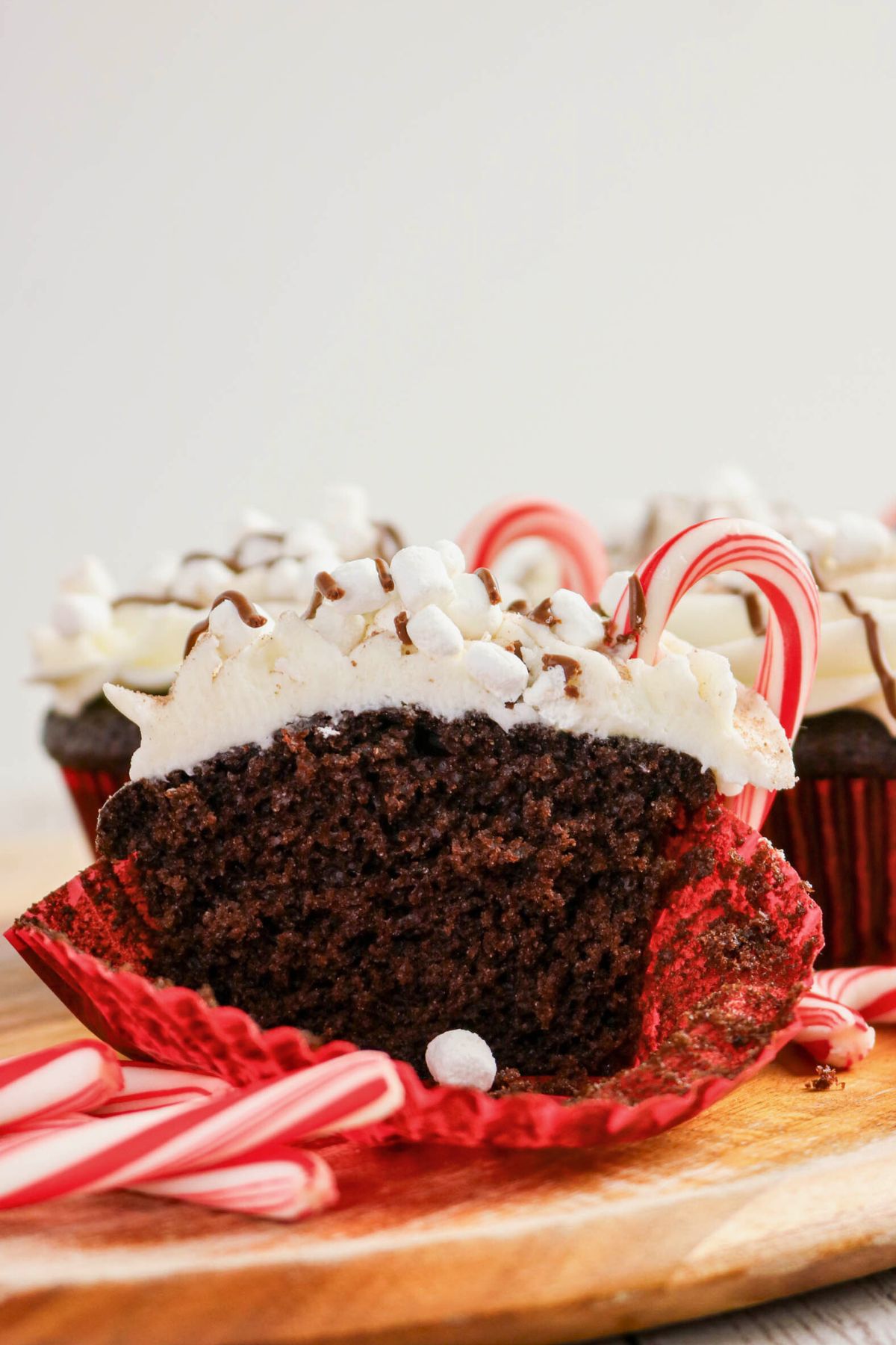 A chocolate cupcake with white frosting, chocolate drizzle, mini marshmallows, and a small candy cane, with the cupcake cut in half on a wooden surface.