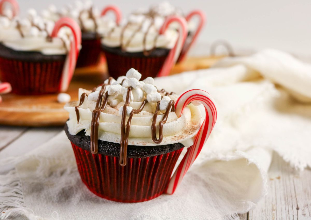 Chocolate cupcakes with white frosting, topped with mini marshmallows and chocolate drizzle, each decorated with a small candy cane shaped like a handle.