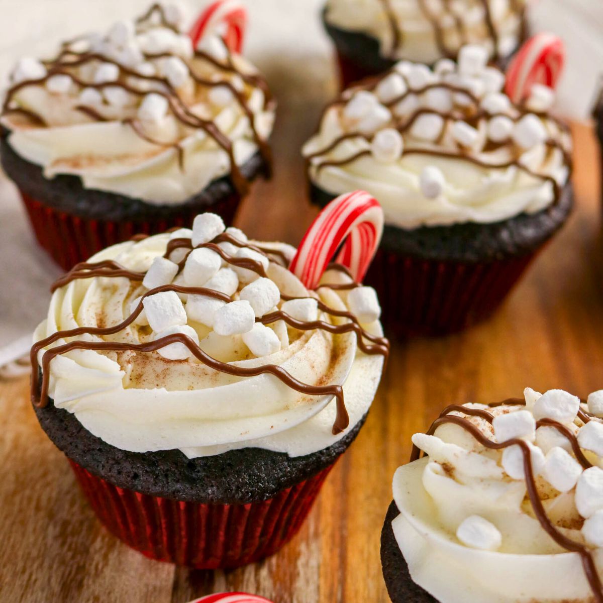 Chocolate cupcakes with white frosting, mini marshmallows, chocolate drizzle, and mini candy canes on top, arranged on a wooden surface.