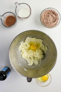Mixing bowl with butter, sugar, and an egg, surrounded by bowls of dry ingredients, milk, cocoa mixture, vanilla extract, and another egg on a white surface.