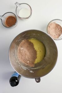 A mixing bowl with cake batter and cocoa powder, surrounded by a measuring cup of milk, a bowl of dry mix, a cup of cocoa liquid, and a bottle of vanilla extract on a white surface.