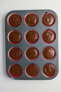 A muffin tin with twelve cups filled with chocolate batter in red paper liners, ready to be baked.