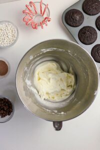 A metal bowl with white frosting is surrounded by chocolate cupcakes, mini chocolate chips, candy canes, cocoa powder, and white sprinkles on a white surface.