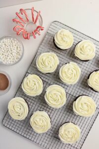A cooling rack with twelve cupcakes topped with white swirled frosting is next to bowls of mini marshmallows, cocoa powder, and striped candy canes on a white surface.