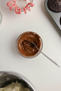 A bowl of chocolate spread with a spoon, surrounded by a muffin tin with baked cupcakes, a bowl of white frosting, and a dish of candy canes on a white surface.