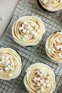 Chocolate cupcakes with swirled white frosting, dusted with cocoa powder, and topped with mini marshmallows on a wire cooling rack.