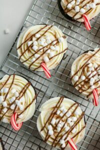 Cupcakes with white frosting, chocolate drizzle, mini marshmallows, and small candy cane handles are arranged on a metal cooling rack.