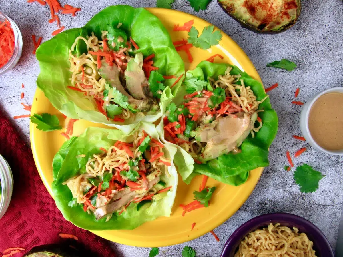 Three lettuce wraps filled with noodles, shredded carrots, chicken, and cilantro are arranged on a yellow plate, with extra vegetables and sauce nearby.
