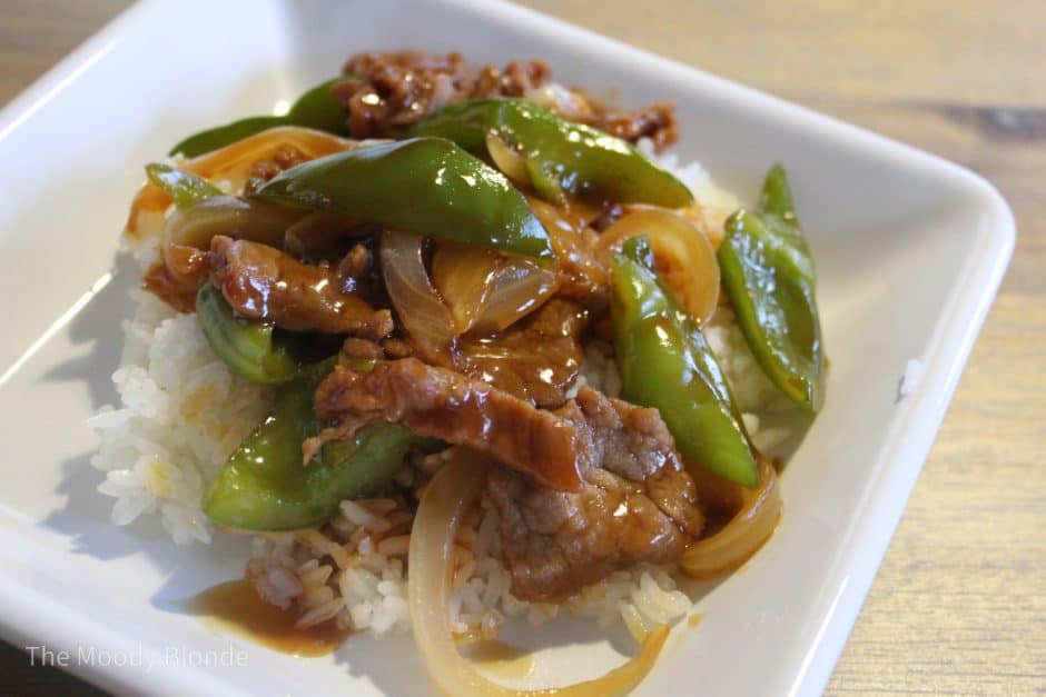 A square white plate with beef stir-fry, green bell peppers, onions, and brown sauce served over white rice.
