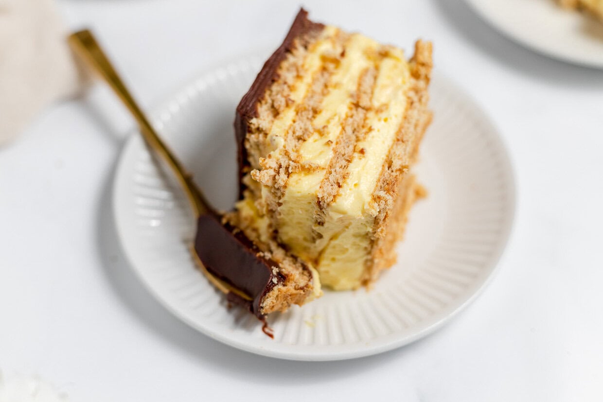 A slice of layered cake with yellow filling and chocolate frosting on a white plate with a gold fork.