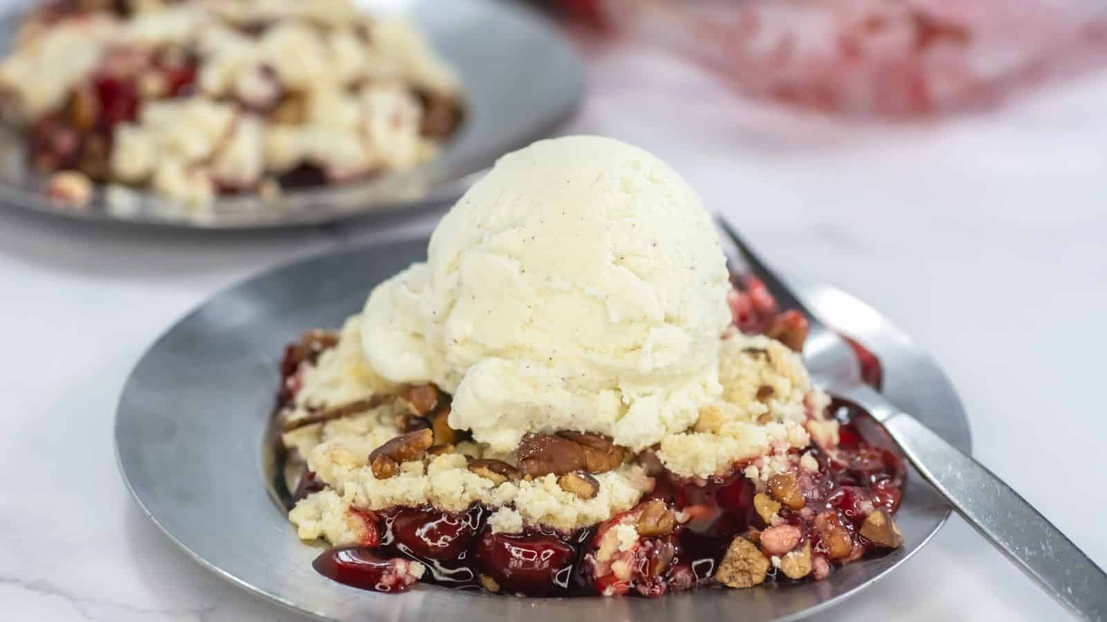 A metal plate with cherry crumble topped with a scoop of vanilla ice cream; a fork is next to the dessert. Another plate is blurred in the background.
