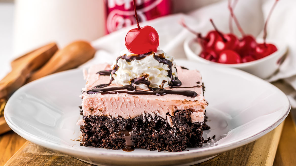 A slice of chocolate and pink frosted cake topped with whipped cream, chocolate drizzle, and a cherry, served on a white plate. A bowl of cherries is in the background.
