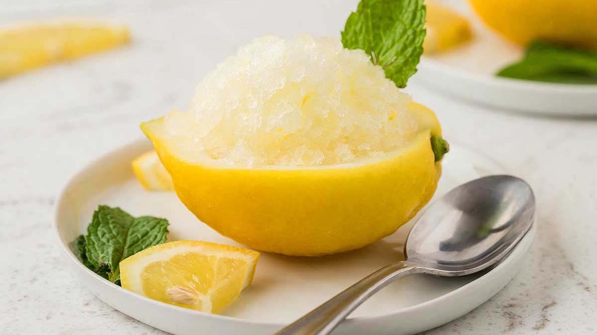 A hollowed lemon half filled with lemon granita, garnished with a mint leaf, sits on a white plate with lemon wedges, mint, and a spoon.