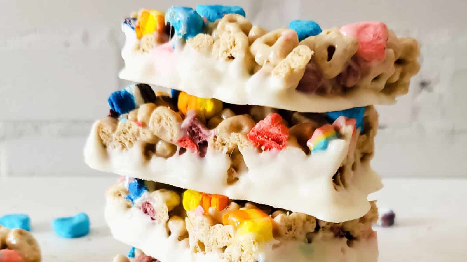 Three stacked cereal bars with colorful marshmallows and coated in white chocolate, set against a light background.