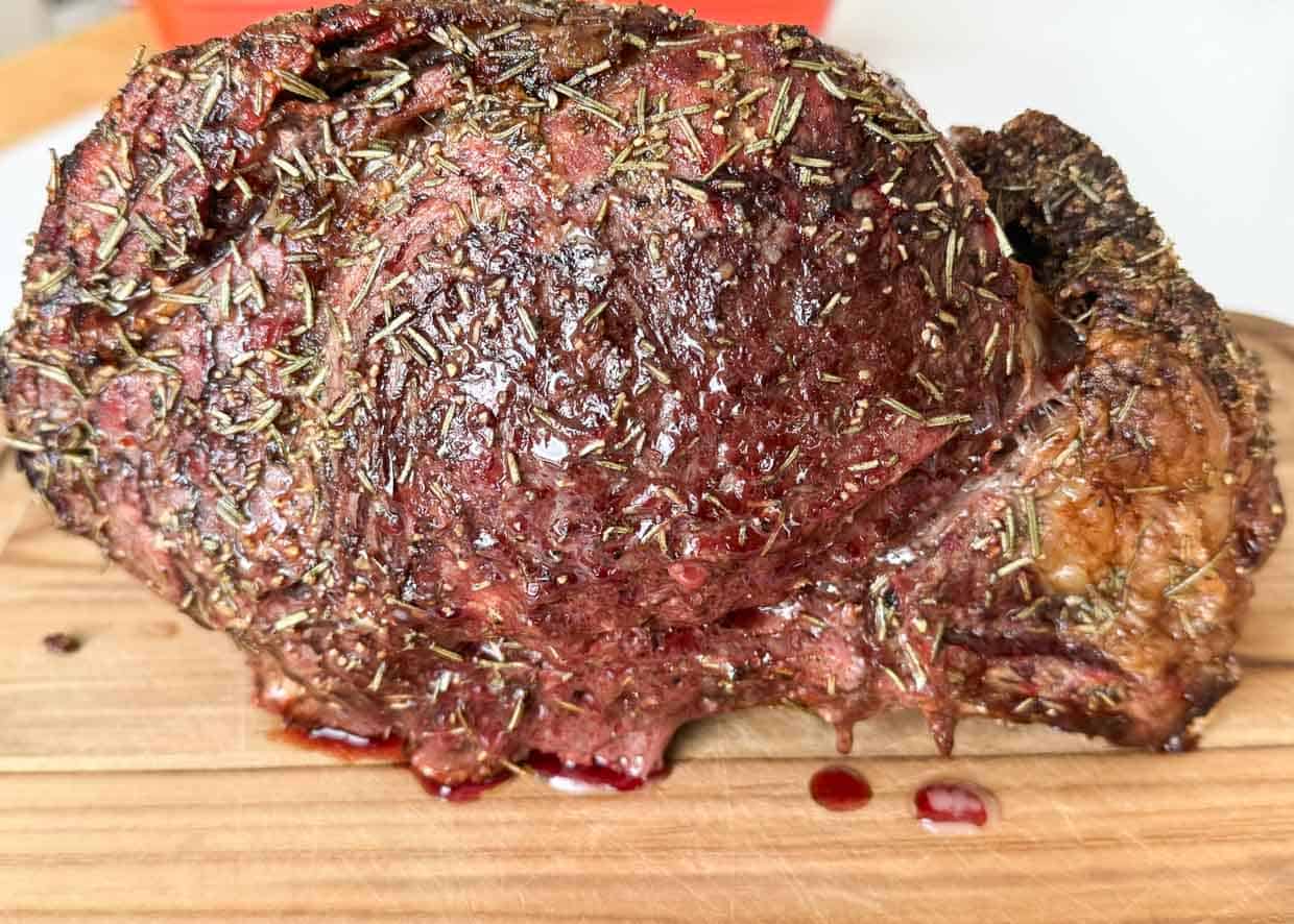 A cooked, herb-crusted roast sits on a wooden cutting board with visible meat juices pooling beside it.
