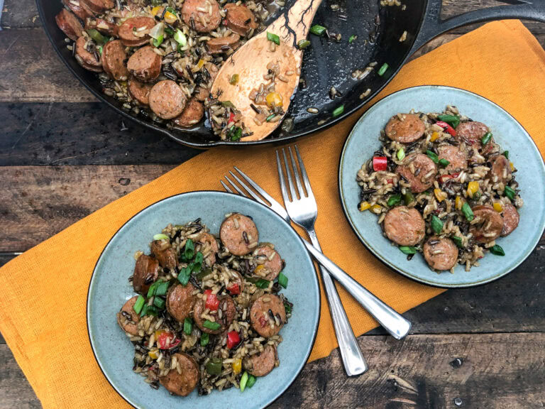 Two plates of sausage and vegetable rice are served next to a skillet with more of the dish. Two forks are on the plates, and the food is set on a wooden table with an orange cloth.