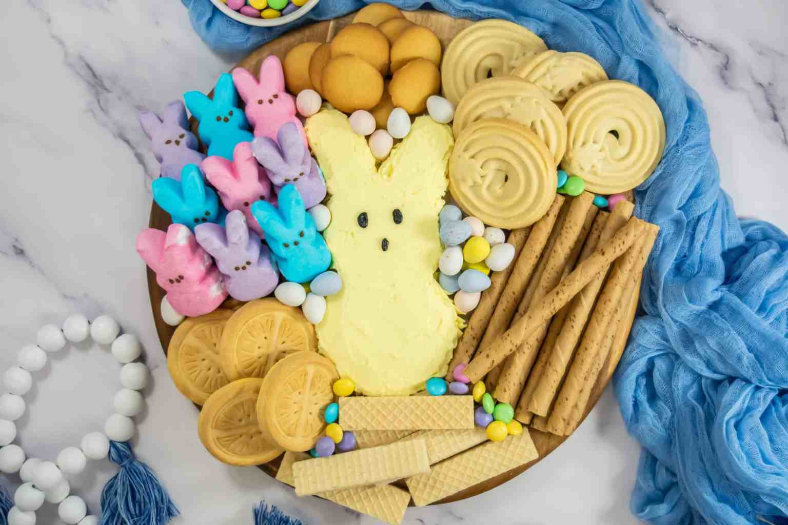 A dessert platter features cookies, wafer rolls, candy eggs, and marshmallow bunny Peeps arranged around a large yellow bunny-shaped dessert. Blue fabric and beads are nearby.