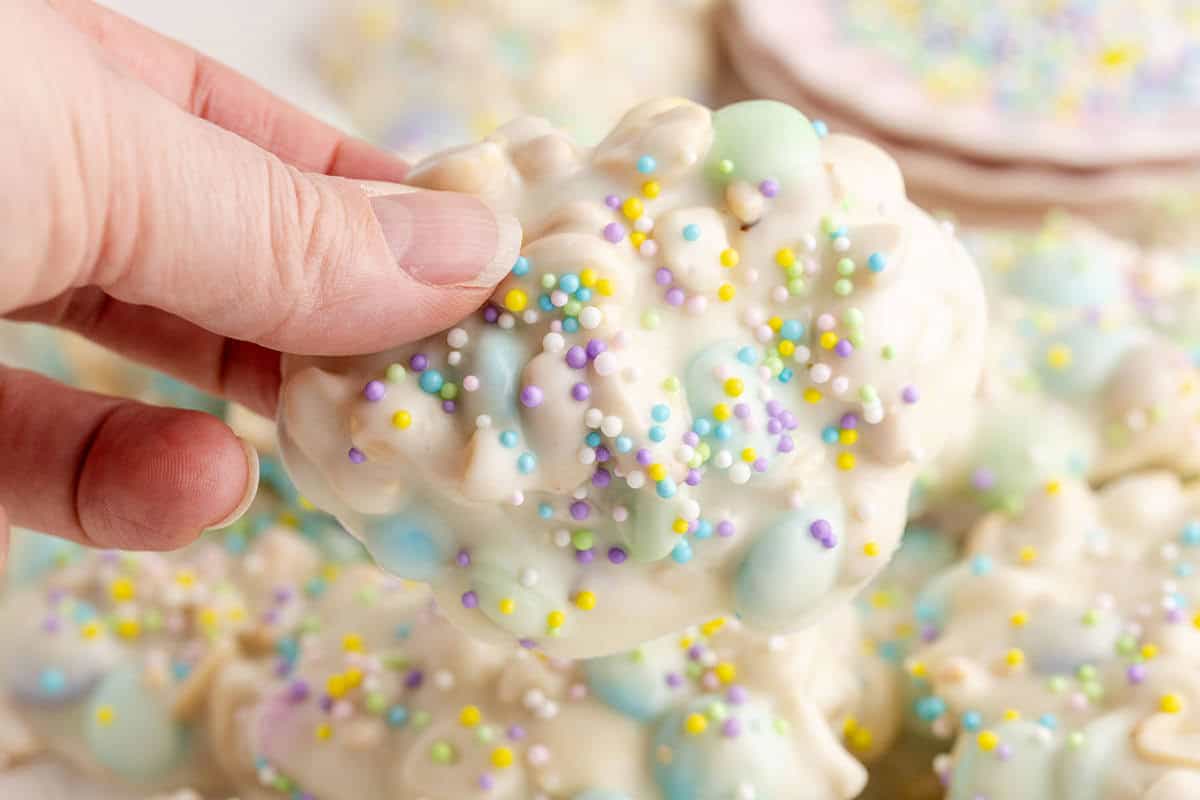 A hand holds a white chocolate cluster with colorful sprinkles and pastel candies on top.