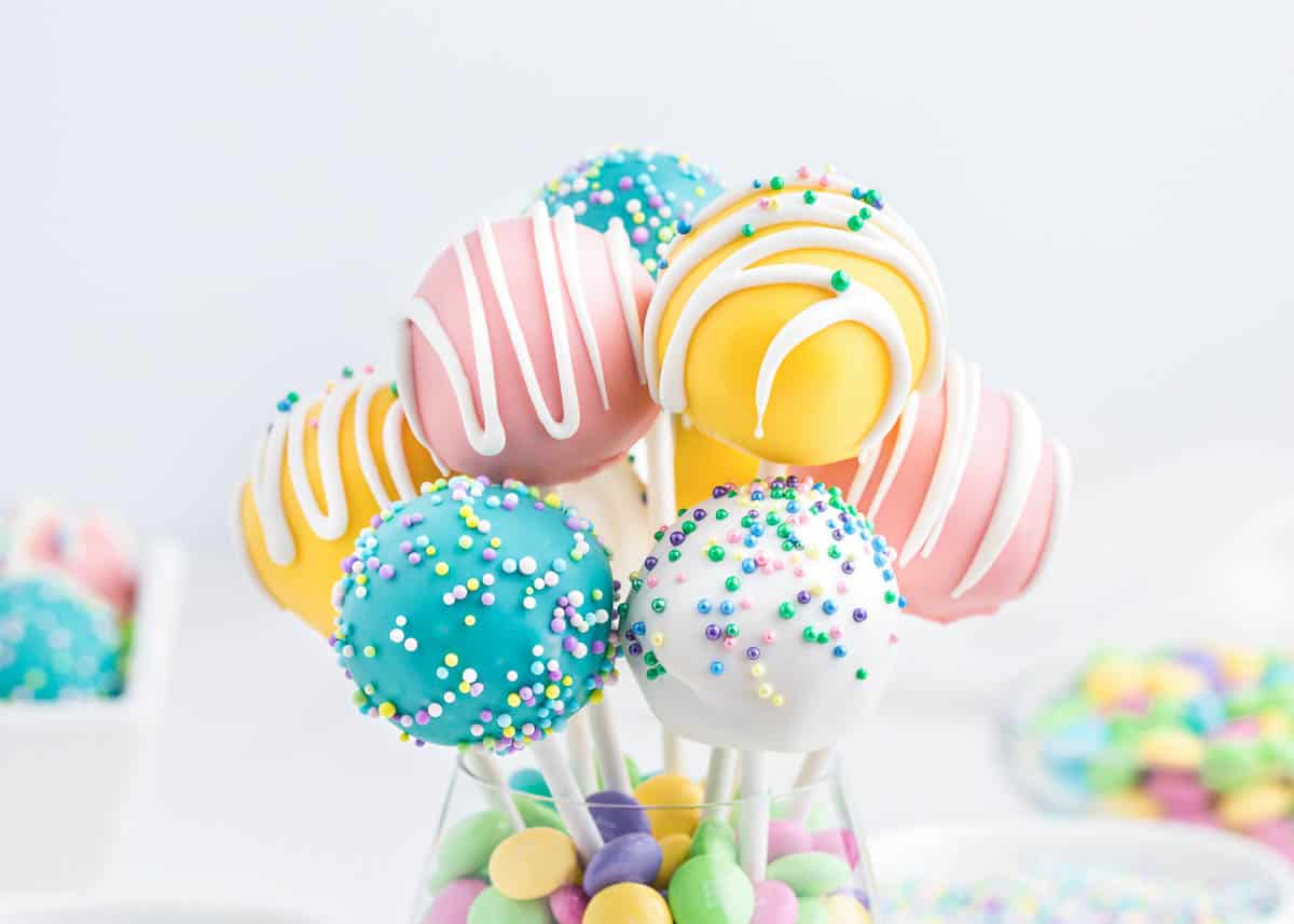 A glass holds colorful cake pops coated in icing and sprinkles, with pastel candies at the base; more cake pops and candies are blurred in the background.