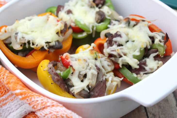 Four halved bell peppers, filled with sliced steak, onions, mushrooms, and topped with melted cheese, arranged in a white baking dish.