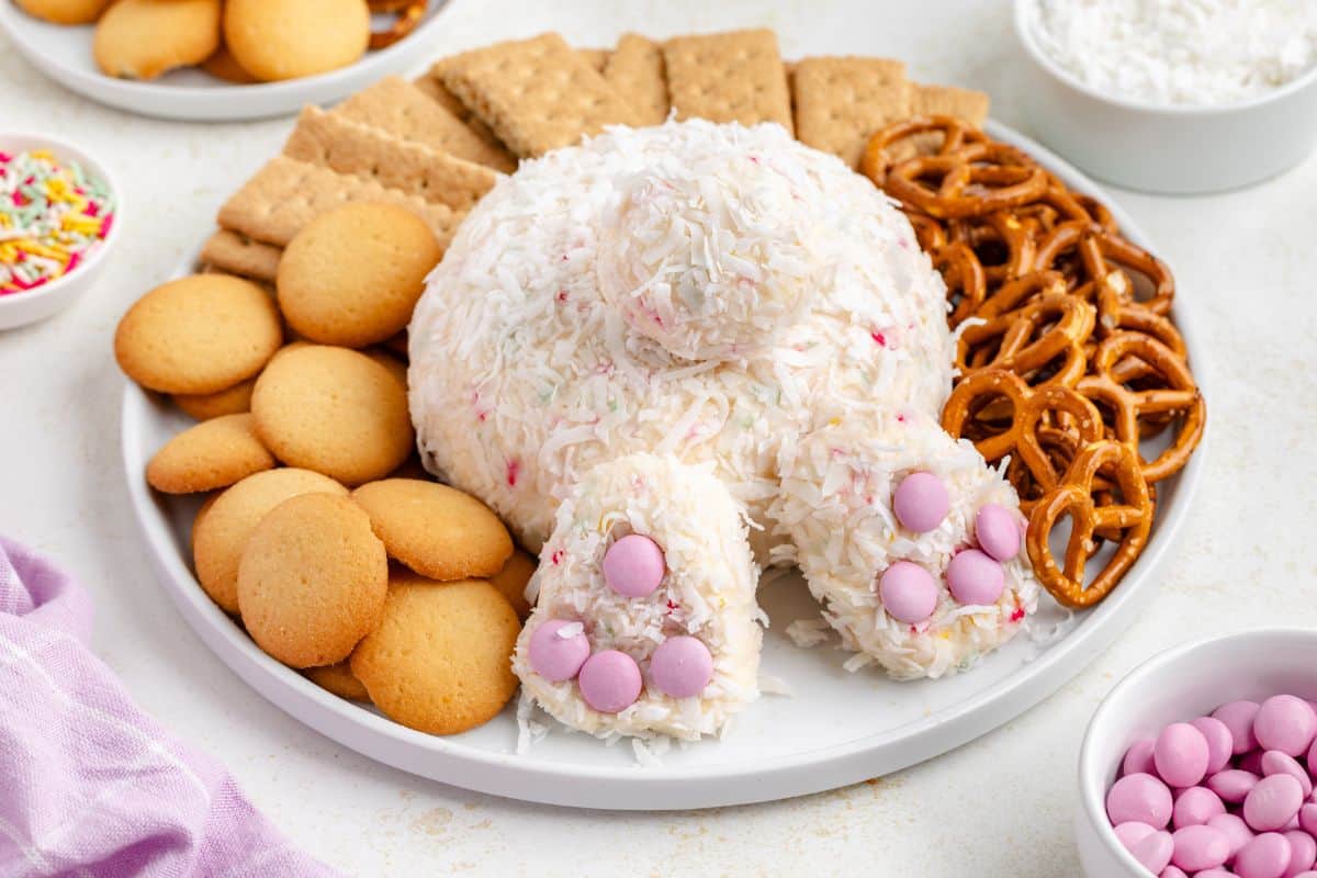 A white bunny-shaped cheese ball covered in shredded coconut is served on a plate with vanilla wafers, graham crackers, and pretzels. Pink candies decorate the bunnys feet.