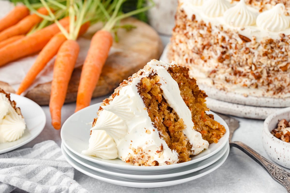 A slice of carrot cake with cream cheese frosting on a plate, with whole carrots and the rest of the cake in the background.
