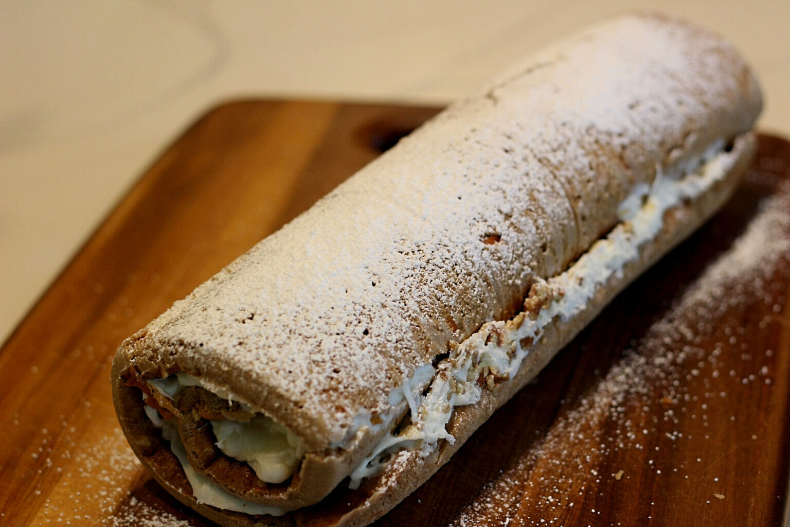 A rolled dessert filled with cream and dusted with powdered sugar sits on a wooden cutting board.