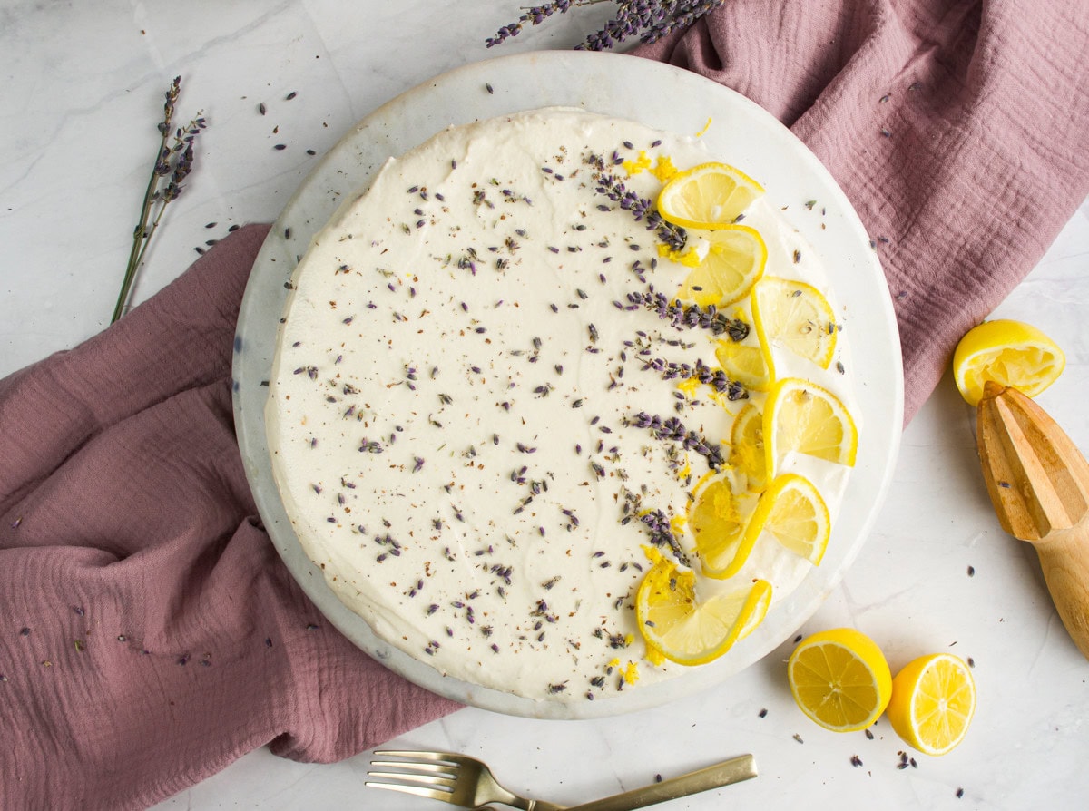 A round cake topped with lemon slices and dried lavender is placed on a marble plate with a mauve cloth, a wooden citrus juicer, and cut lemons nearby.