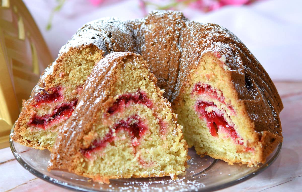 A bundt cake with a swirl of red fruit filling is sliced and arranged on a glass plate, dusted with powdered sugar.