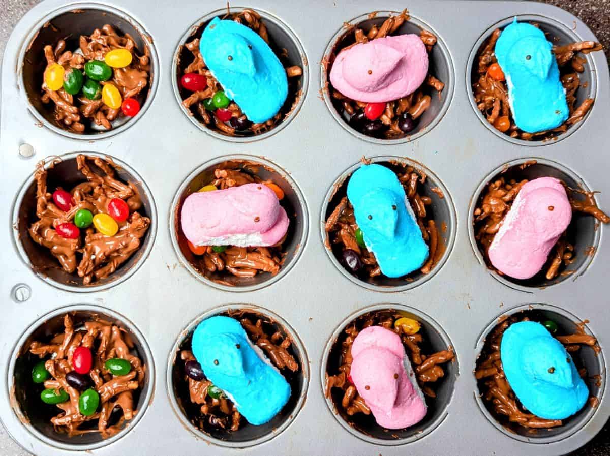 A muffin tin filled with chocolate-coated chow mein noodle nests, some topped with jelly beans, others with pink or blue marshmallow Peeps shaped like chicks.