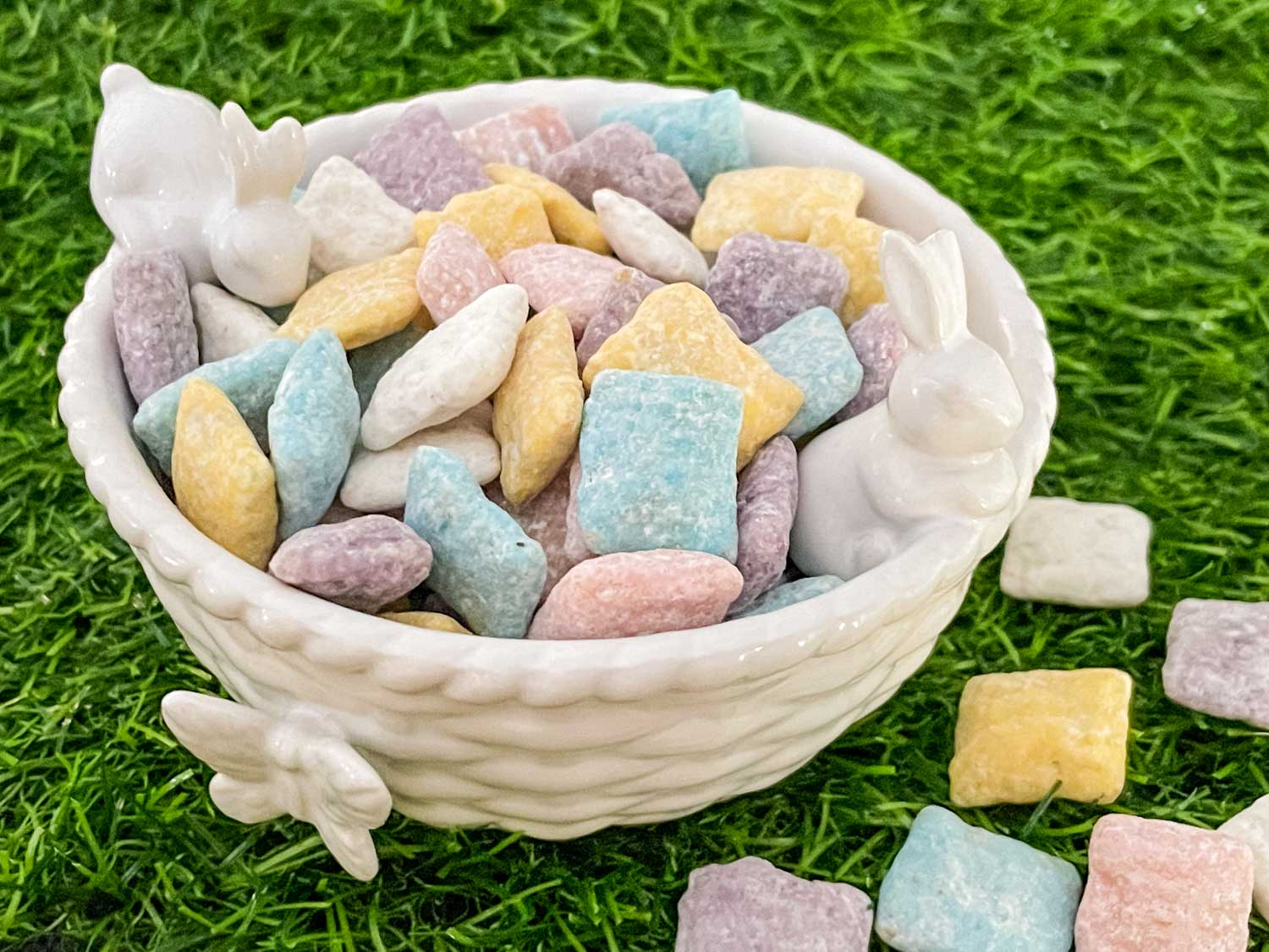 A white ceramic bowl with bunny decorations filled with pastel-colored candy squares, placed on green artificial grass.