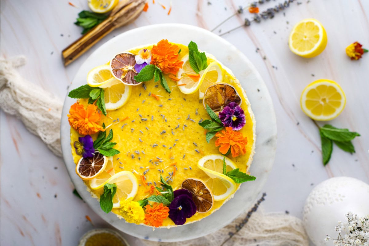 A round yellow cake decorated with lemon slices, colorful edible flowers, mint leaves, and dried citrus, placed on a white marble surface with lemon halves and herbs nearby.