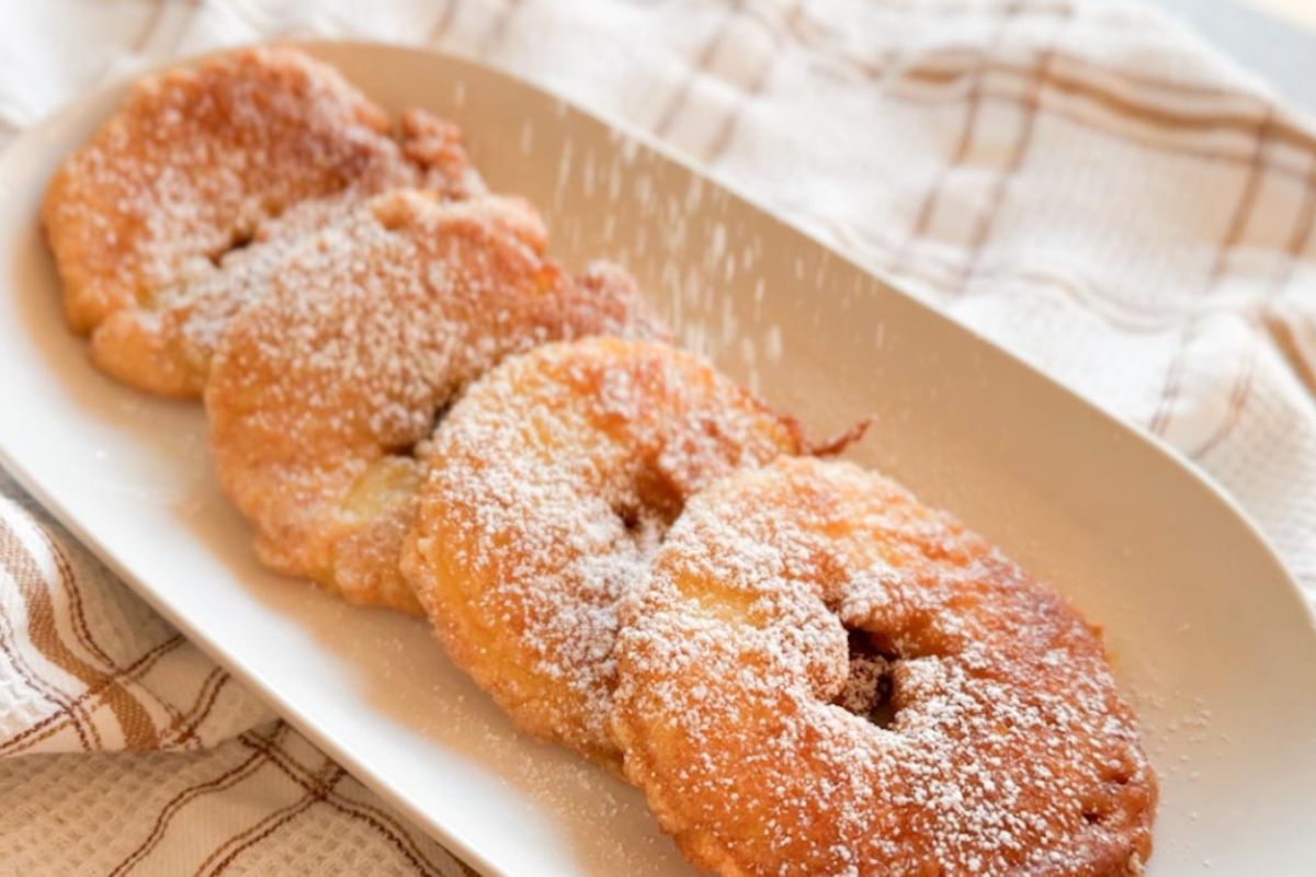 Four round apple fritters dusted with powdered sugar are arranged in a row on a white oval plate, set on a checkered cloth.