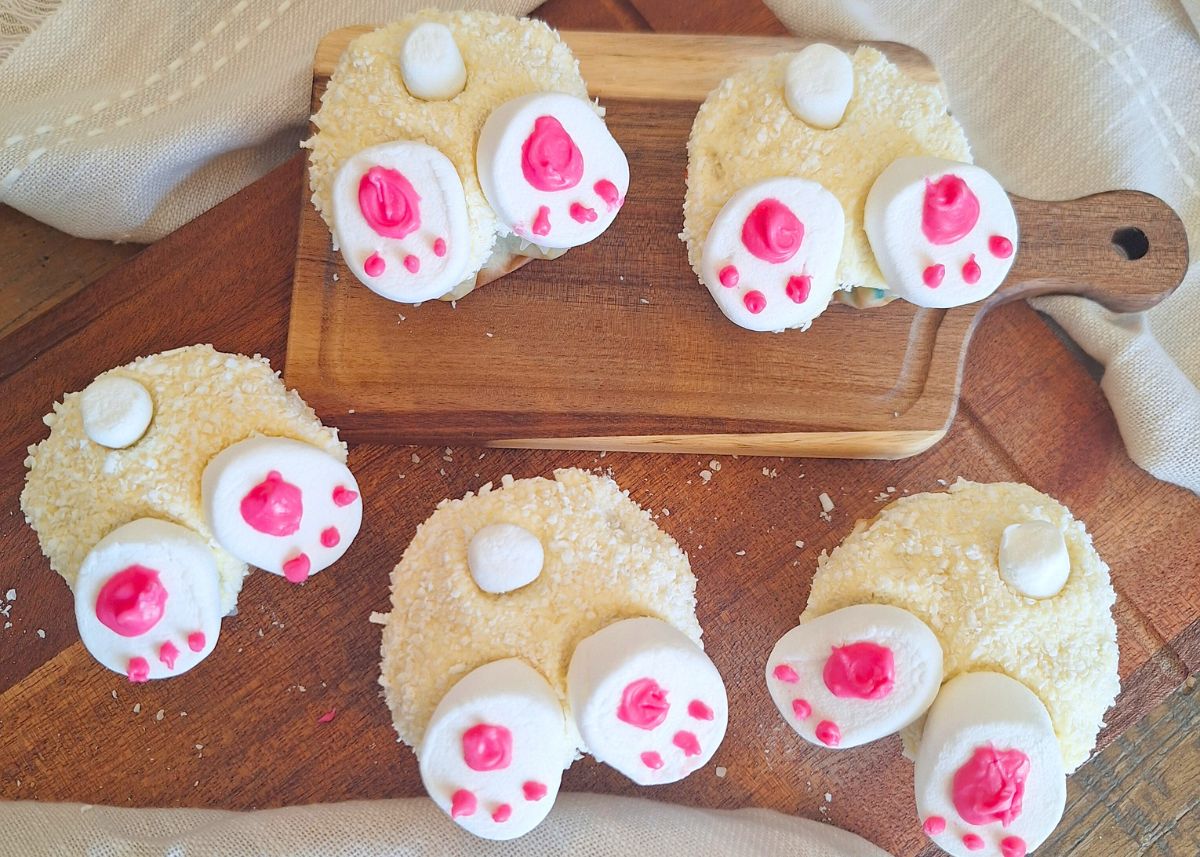 Five round cookies decorated with white and pink icing to resemble bunny feet are arranged on a wooden board and surface.