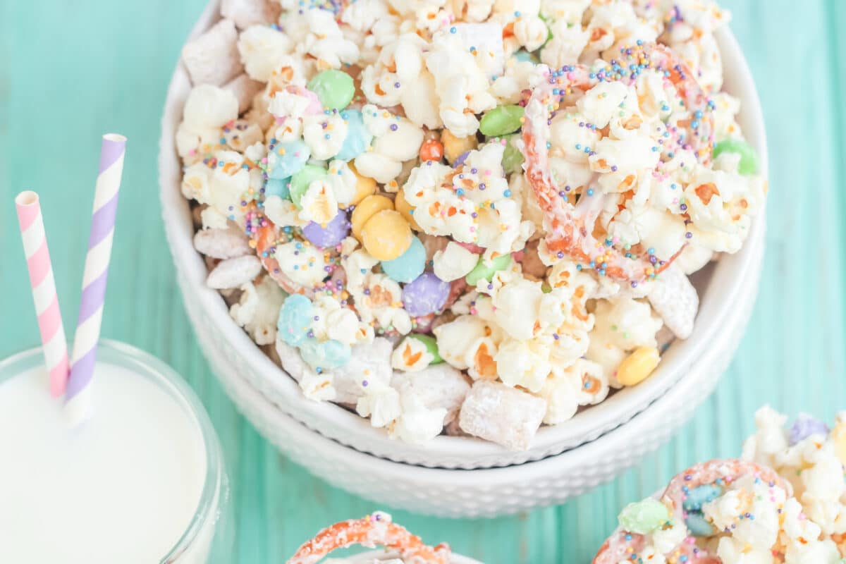 A bowl of popcorn snack mix with colorful candies, sprinkles, and pretzels sits on a light blue wooden surface next to a glass of milk with two striped straws.