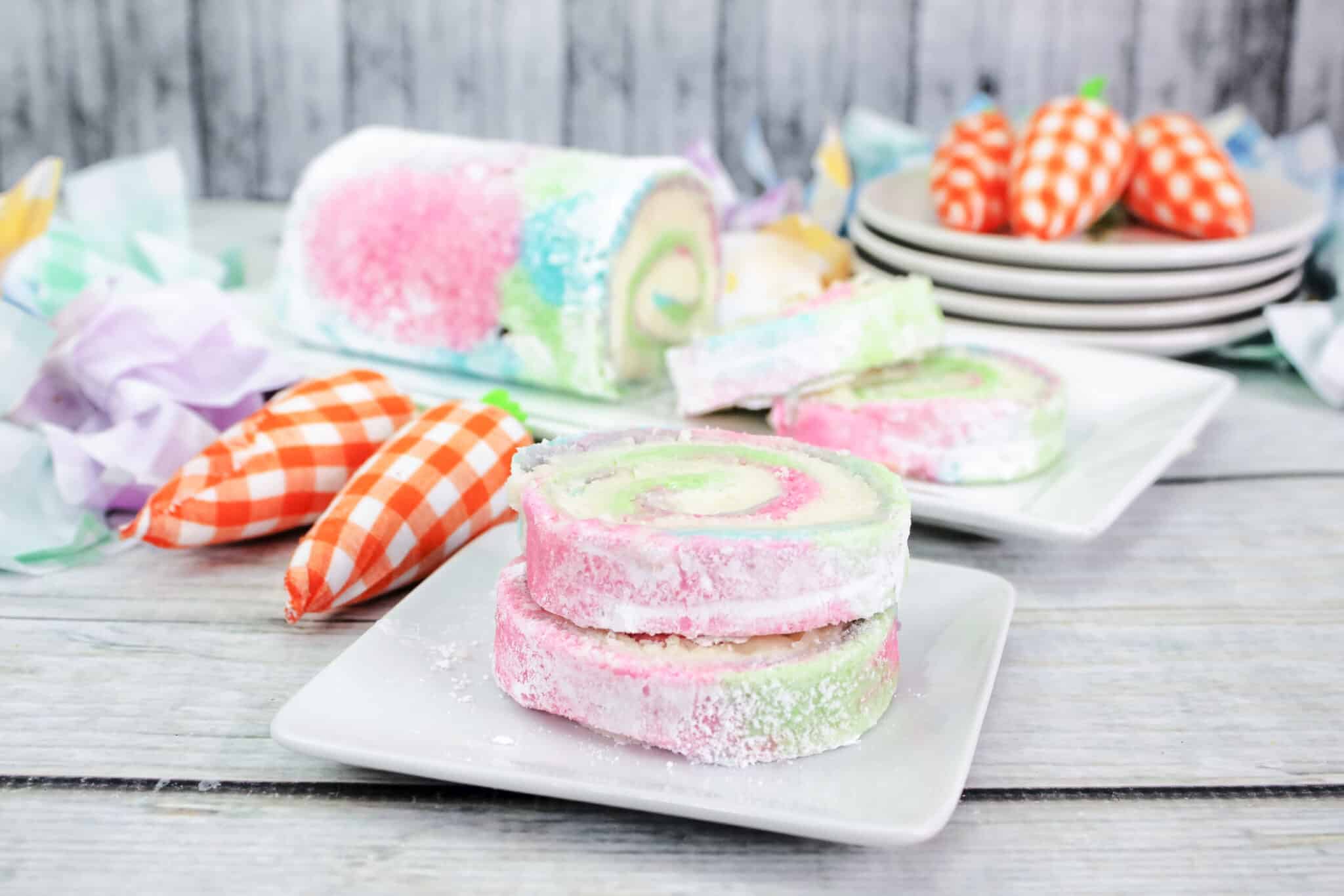 Colorful swirled cake roll slices dusted with powdered sugar are served on white plates, with decorative orange gingham carrots and a whole cake roll in the background.