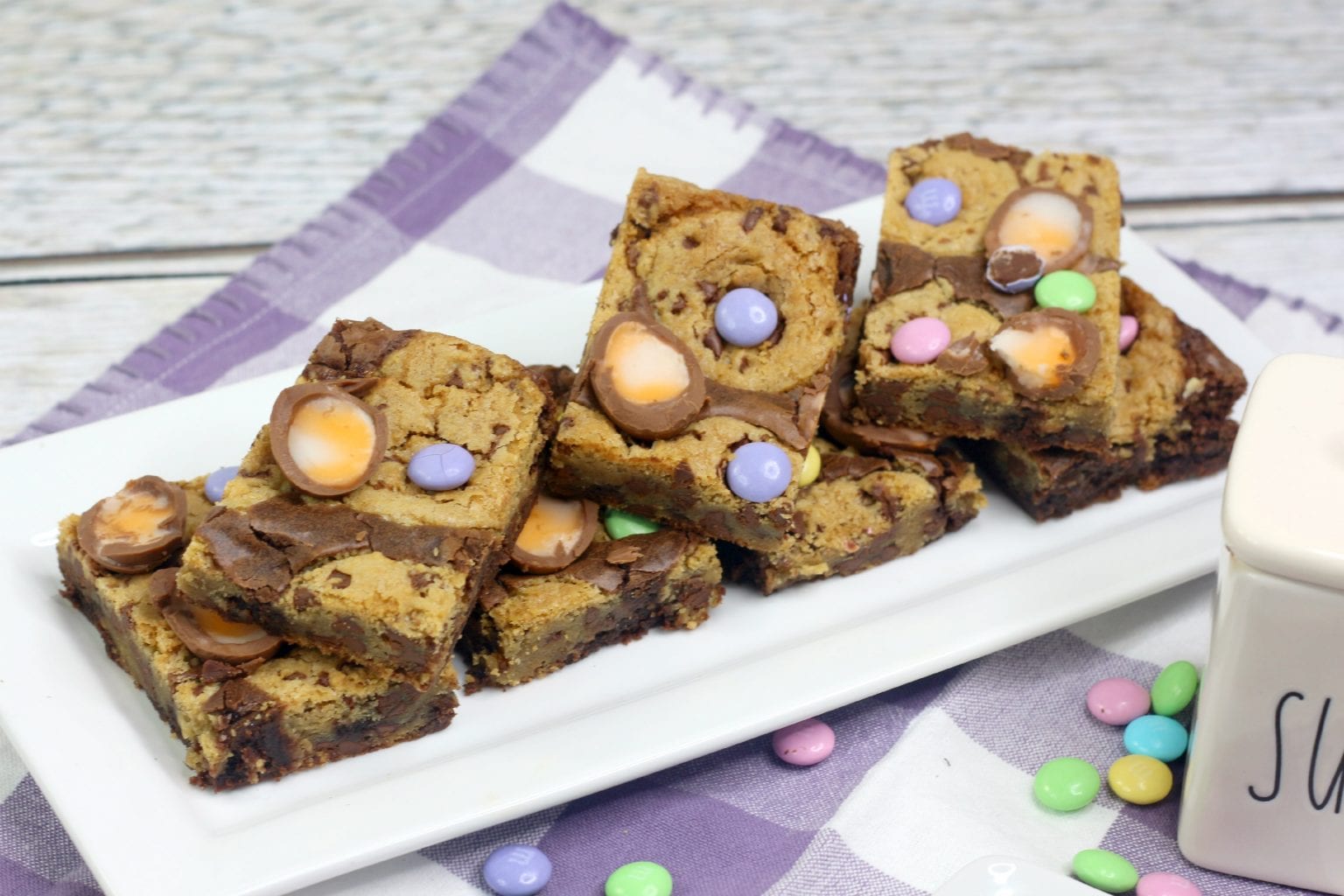A plate of chocolate chip cookie bars topped with colorful candy-coated chocolates, placed on a white and purple checkered cloth.