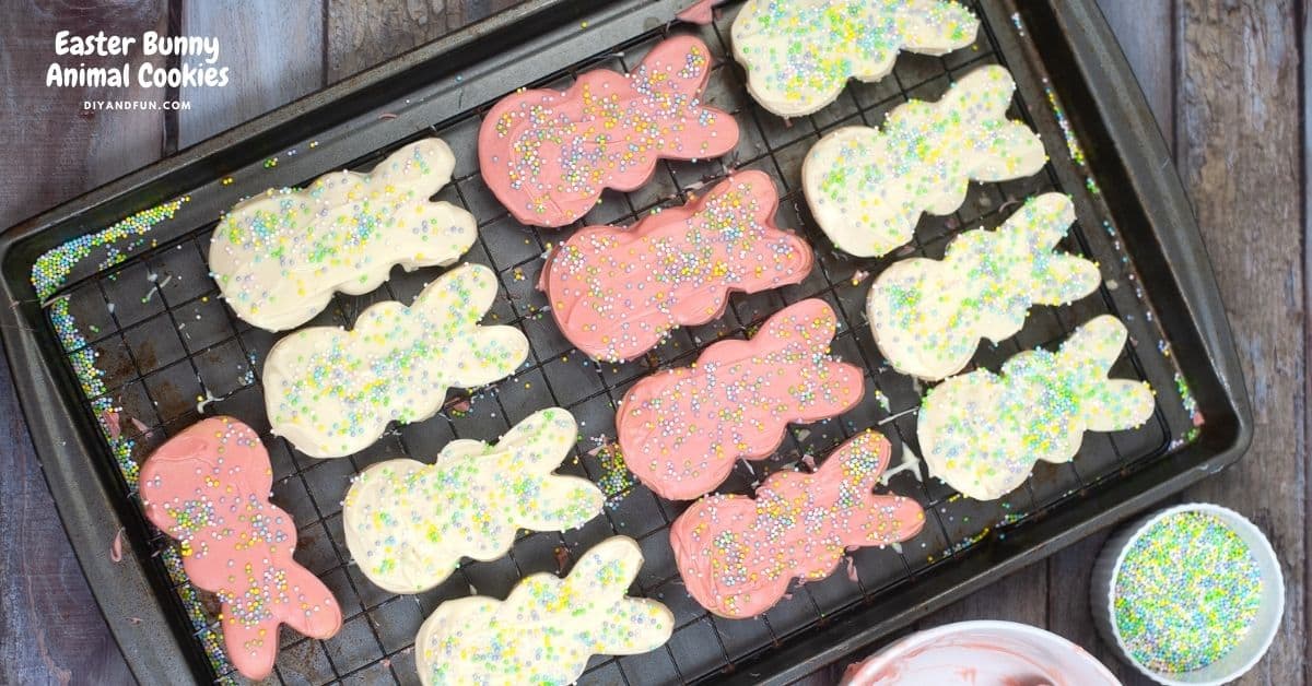 A baking sheet with twelve bunny-shaped cookies, half coated in pink icing and half in white icing, topped with colorful sprinkles. A bowl of sprinkles and frosting are nearby.