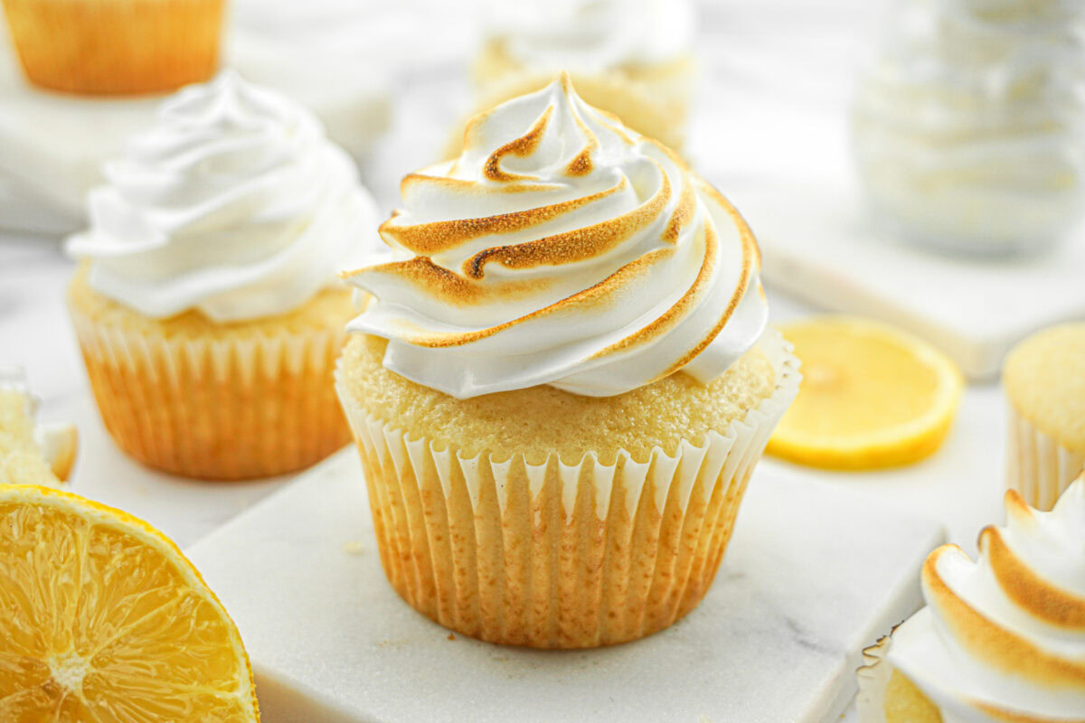 A lemon cupcake with swirled, toasted meringue frosting sits on a marble surface, with lemon slices and more cupcakes in the background.