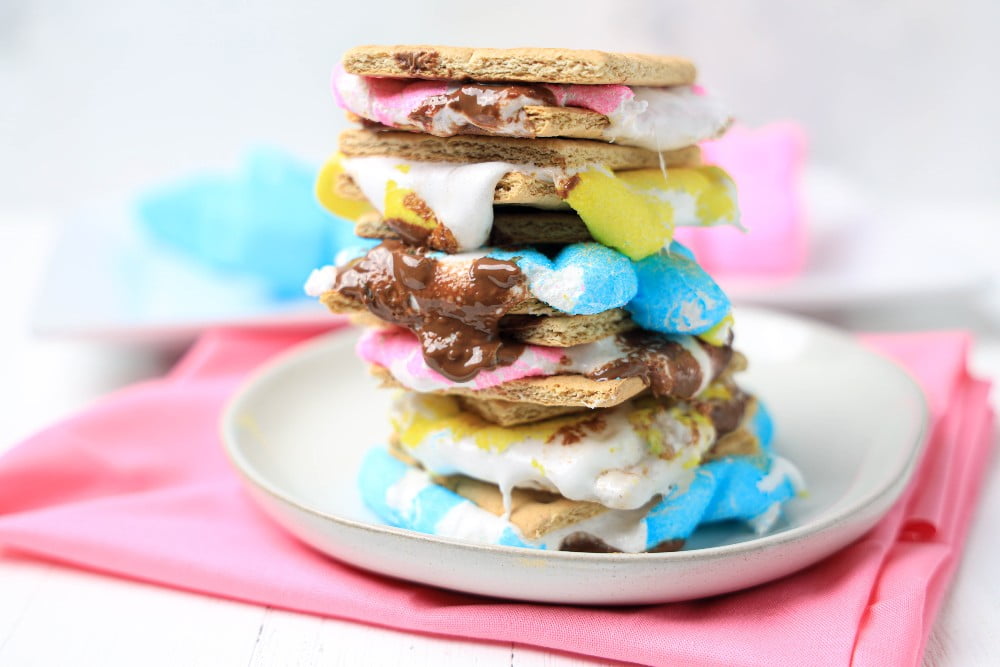 A stack of smores filled with melted chocolate and colorful marshmallow chicks on a white plate, with a pink napkin underneath.