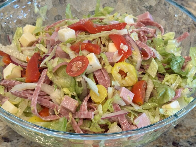 A glass bowl filled with an Italian chopped salad featuring lettuce, tomatoes, salami, cheese cubes, onions, and pepper rings.