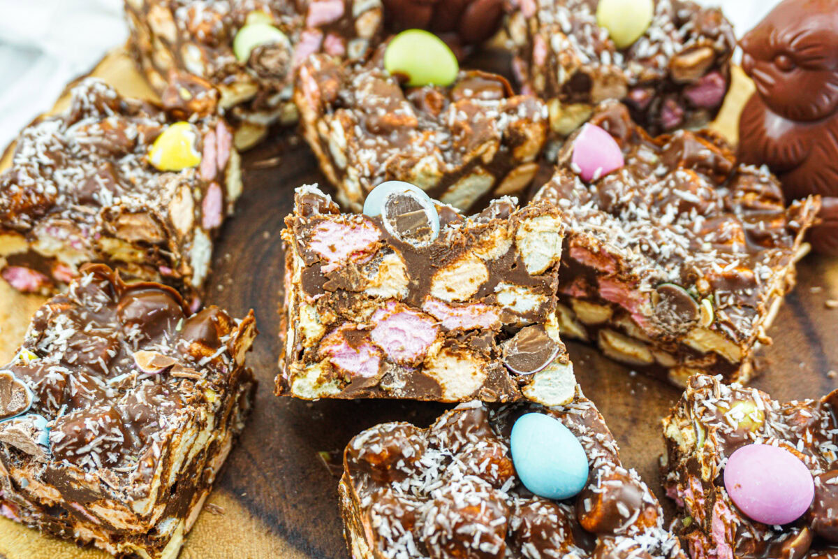 Sliced chocolate rocky road bars with marshmallows, biscuits, and candy eggs are arranged on a wooden board.