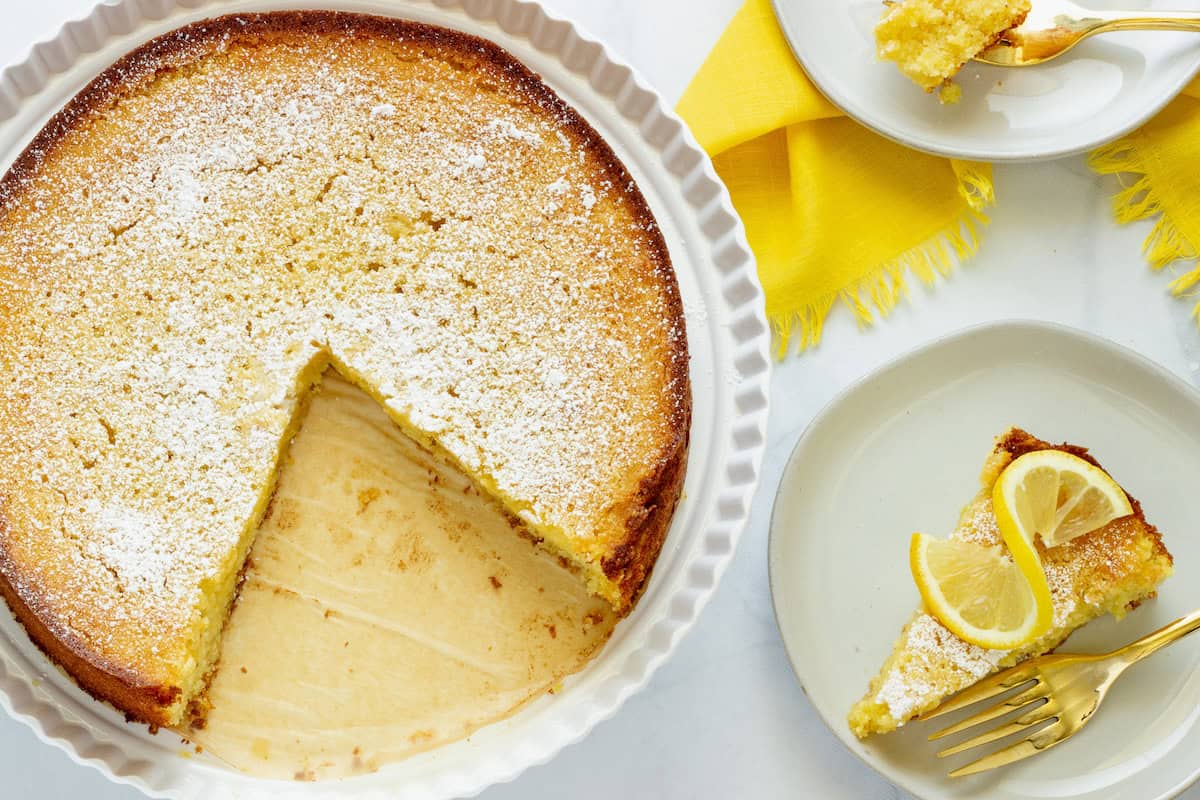 A round lemon cake with a missing slice, dusted with powdered sugar. Two plates each hold a slice, one garnished with lemon wedges. Yellow napkin and gold fork are beside the plates.