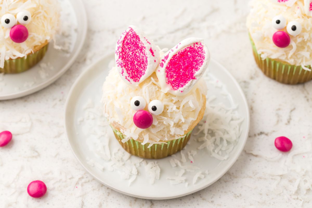 A cupcake decorated to look like a bunny with white frosting, coconut flakes, pink candies for noses, candy eyes, and pink-sprinkled ears on a white plate.