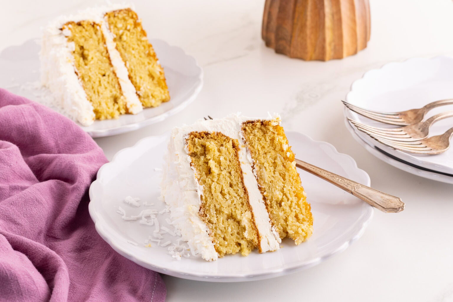 Two slices of layered yellow cake with white frosting and shredded coconut on white plates, one with a fork; a stack of plates and forks in the background.