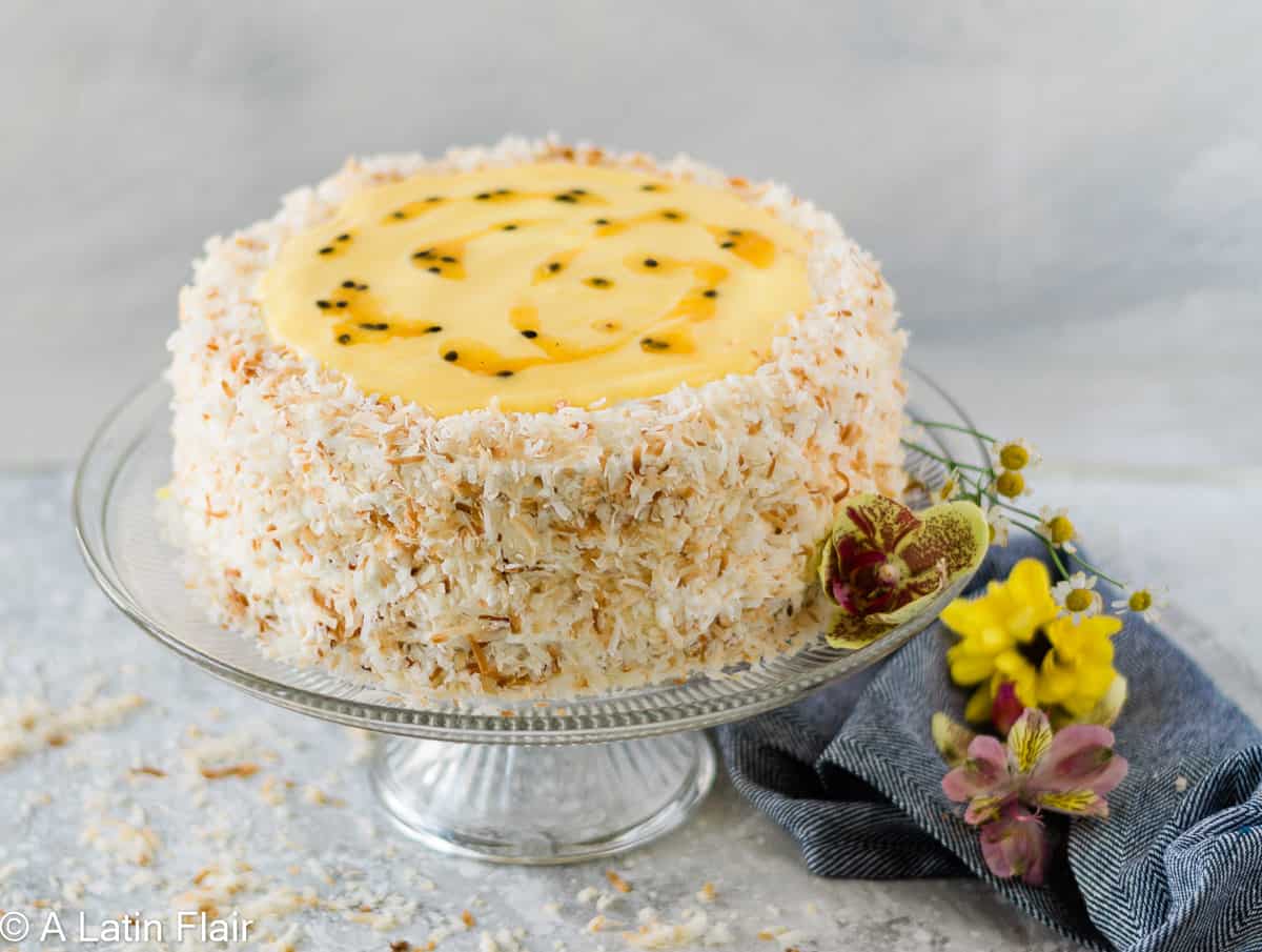A round cake covered in toasted coconut flakes, topped with yellow passion fruit glaze, displayed on a glass cake stand with flowers and a dark cloth nearby.