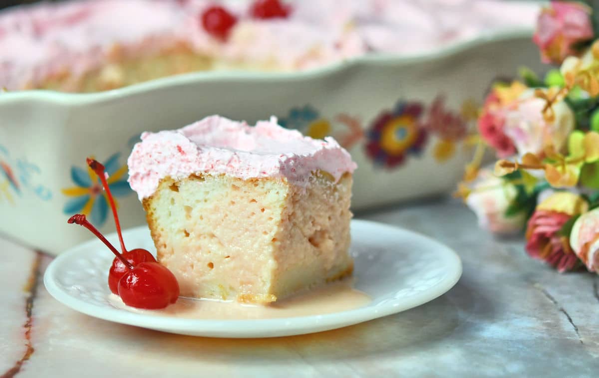 A slice of pink-frosted cake on a white plate with two maraschino cherries, with a floral baking dish and flowers in the background.