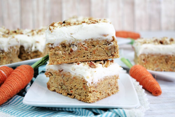 Two frosted carrot cake bars topped with chopped nuts are stacked on a white plate, with decorative carrots and more cake bars in the background.