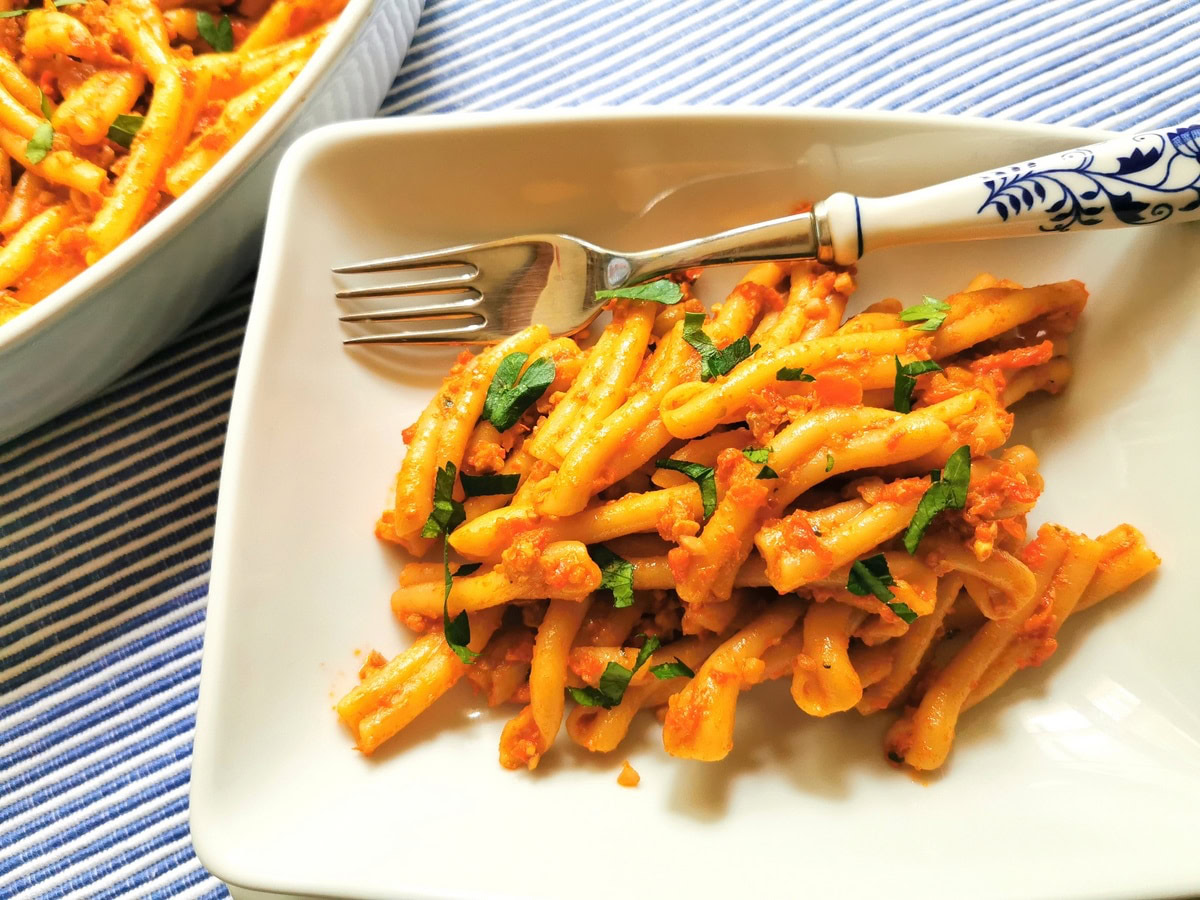 A plate of pasta with tomato-based sauce and chopped herbs, served with a fork on a white rectangular dish on a striped tablecloth.